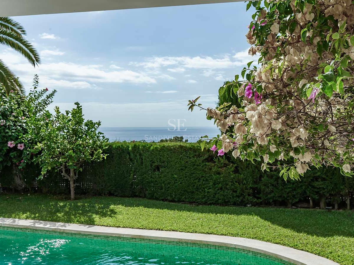 Piscina turquesa rodeada de césped verde y flores con vistas panorámicas al mar Mediterráneo.