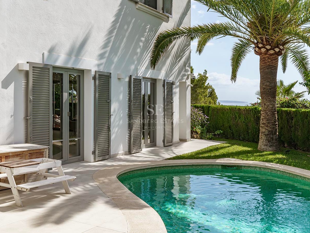 Piscina turquesa junto a una villa blanca con una palmera, persianas grises y vistas lejanas al mar.