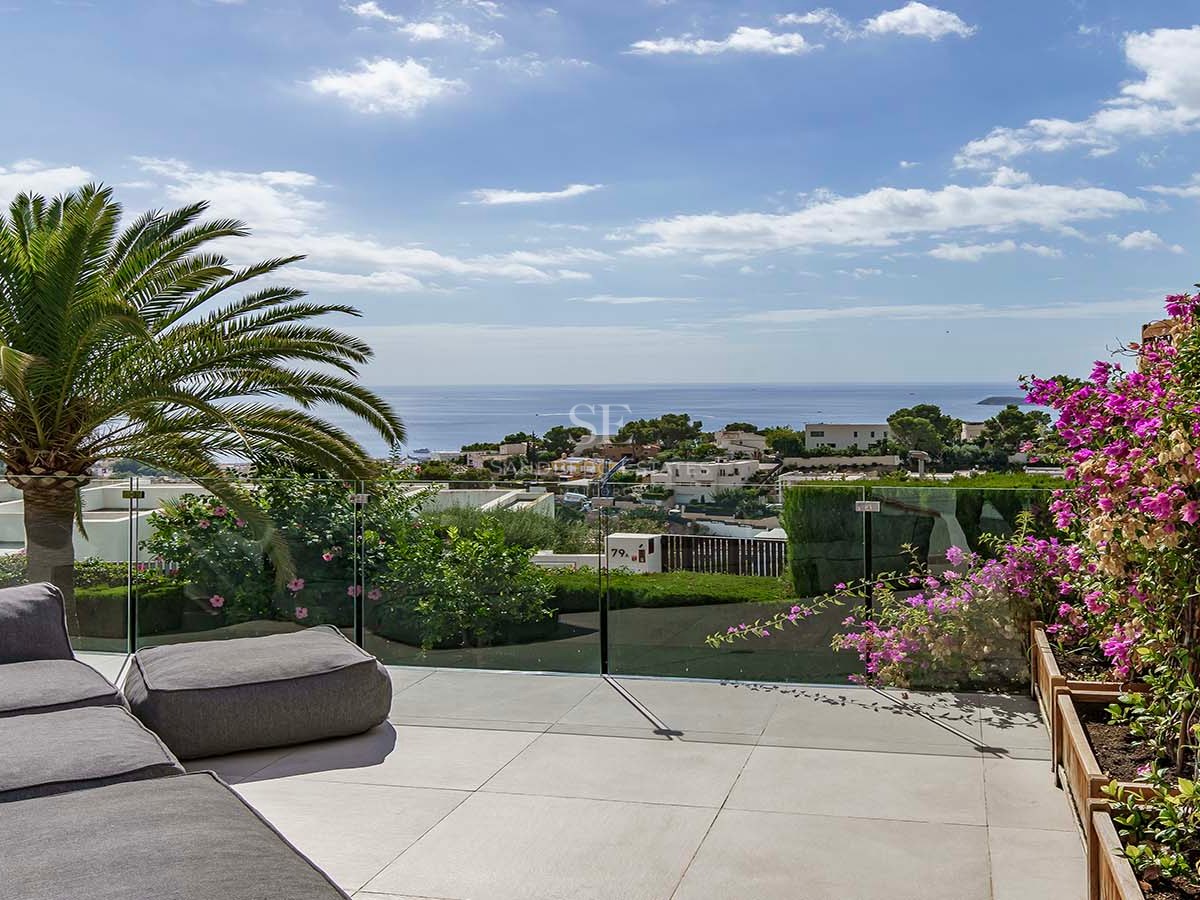 Terraza de lujo con sofá gris, barandilla de cristal, palmeras y vistas impresionantes al mar y al pueblo.