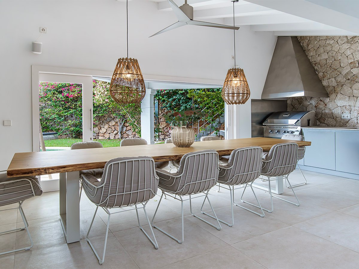 Mesa de madera maciza con sillas modernas y lámparas de mimbre junto a una cocina exterior con barbacoa de acero.