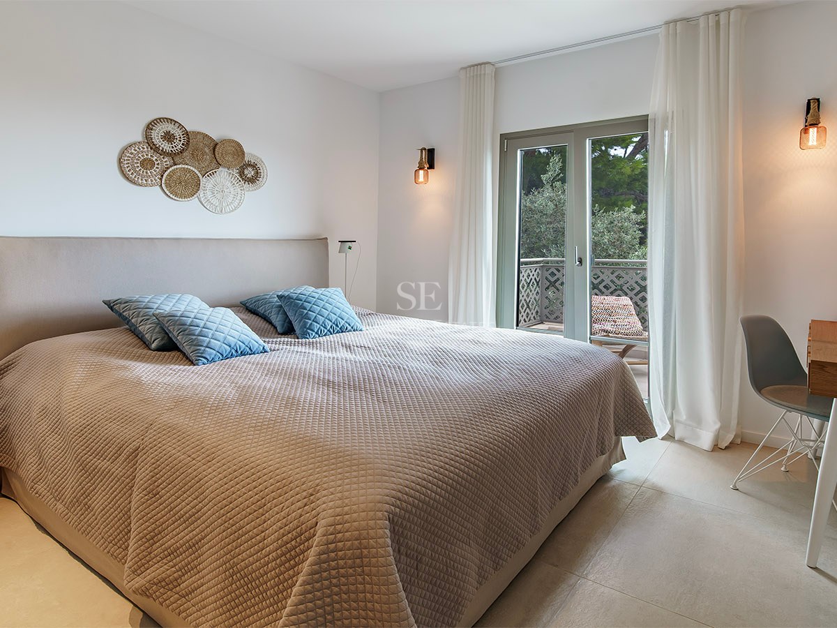 Dormitorio moderno con cama grande, colcha beige, cojines azules, escritorio de madera y puerta de cristal al balcón.