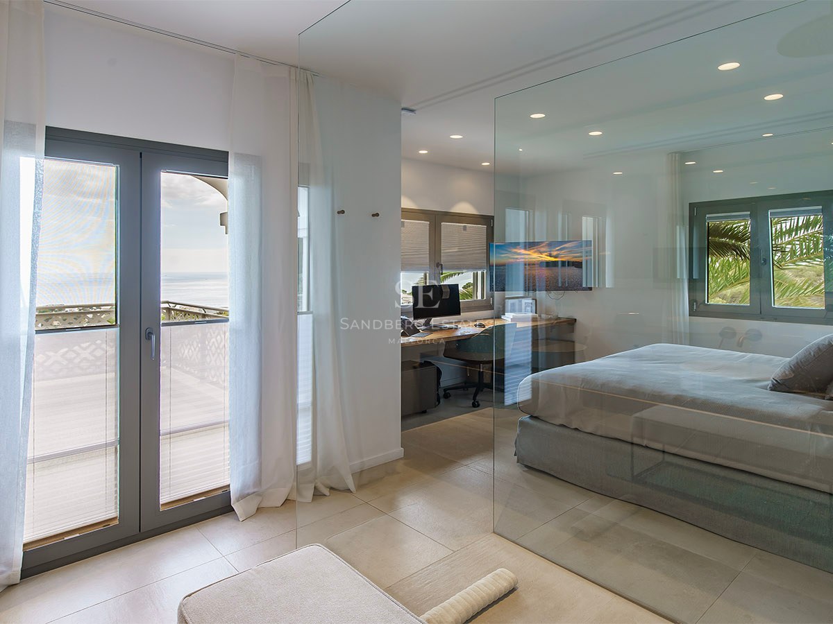 Dormitorio moderno con pared de cristal, zona de oficina integrada y balcón con vistas al mar.