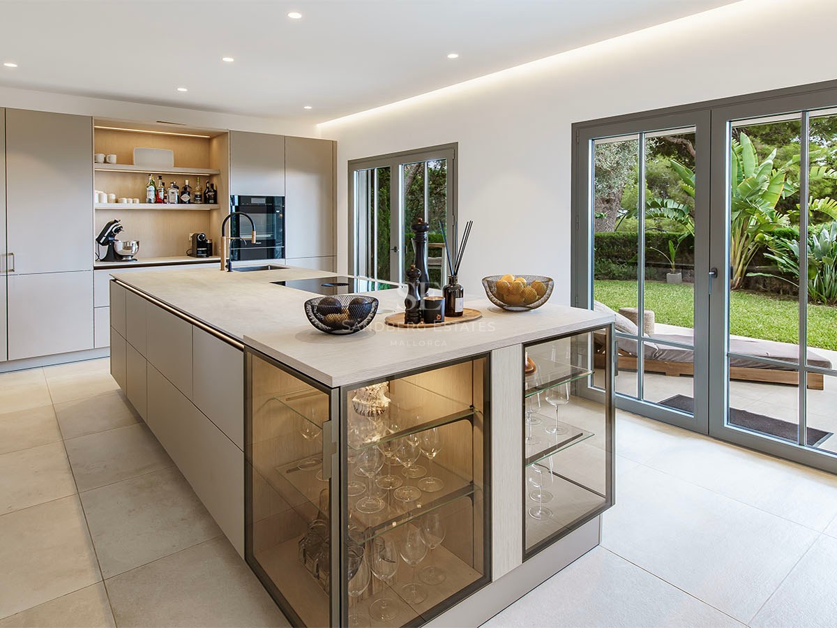 Cocina moderna con isla grande, vitrinas integradas y puertas de cristal que dan a un jardín verde.