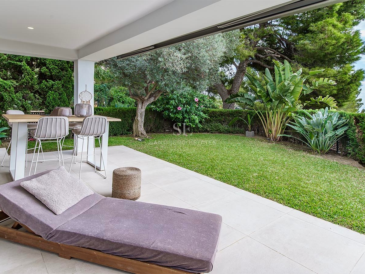 Terraza cubierta con mesa de comedor, tumbona y jardín impecable con vistas parciales al mar.