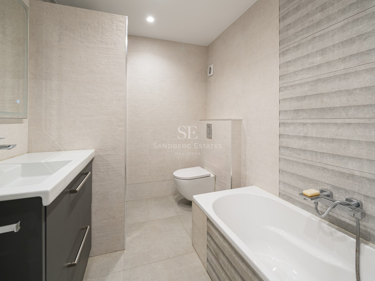 Baño moderno con bañera blanca, inodoro suspendido y tocador gris con azulejos beige texturizados.