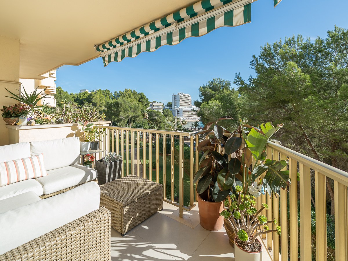 Terraza con sofás de mimbre blanco, plantas verdes y toldo a rayas con vistas a árboles y edificios.