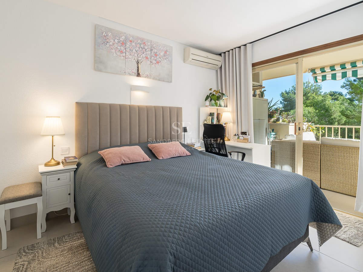 Dormitorio luminoso con cama acolchada gris, escritorio y puertas correderas de cristal que dan a un balcón soleado.