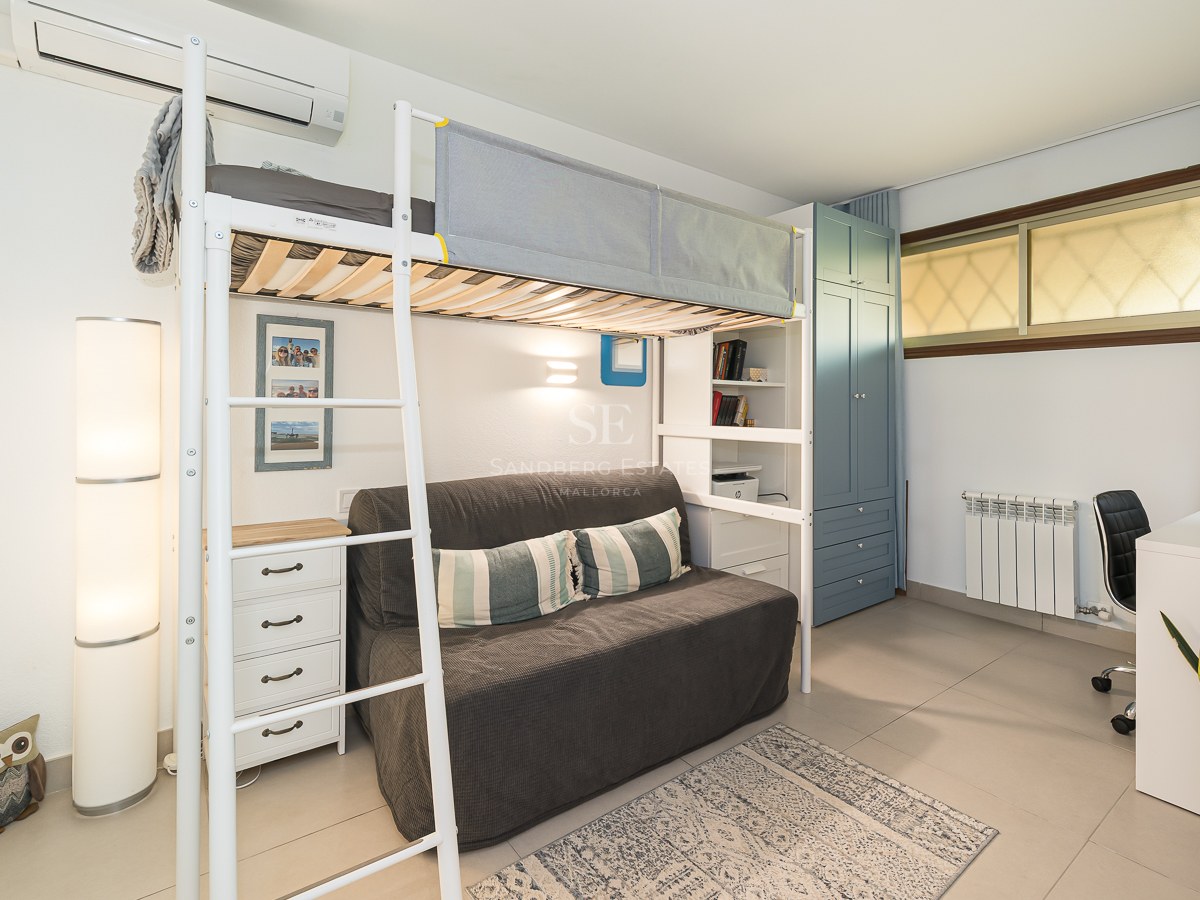 Cama alta de metal blanco sobre un sofá gris, junto a un escritorio y un armario azul en una habitación luminosa.
