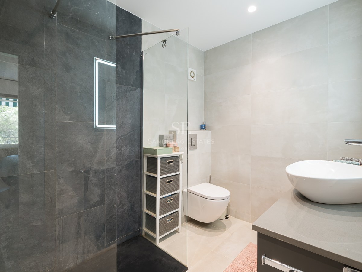 Baño moderno con ducha de cristal, azulejos gris oscuro, lavabo sobre encimera blanco y sanitario suspendido.