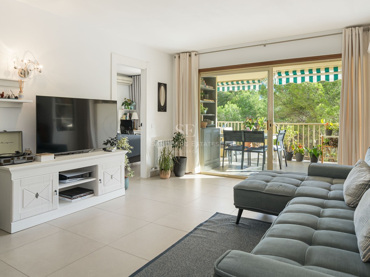 Salón amplio con sofá gris, televisión sobre mueble blanco y puertas correderas de cristal que dan a una terraza verde.