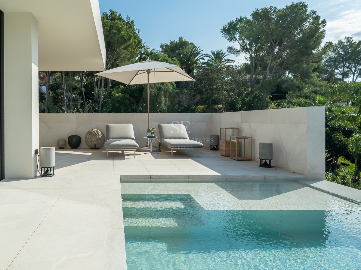 Piscine à débordement moderne avec terrasse en pierre blanche, chaises longues, parasol et arrière-plan de verdure.