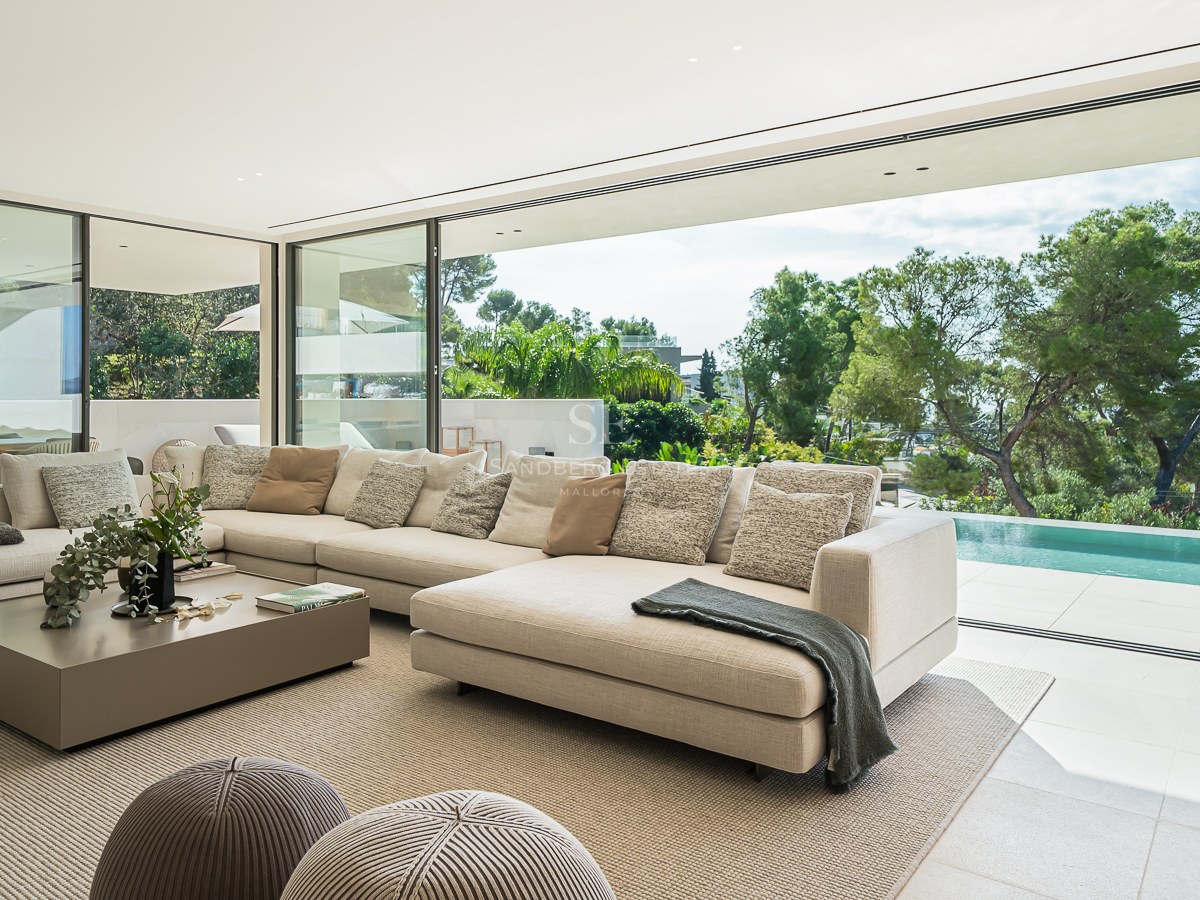 Un salon moderne et lumineux avec un grand canapé d'angle crème donnant sur une piscine privée et des arbres.