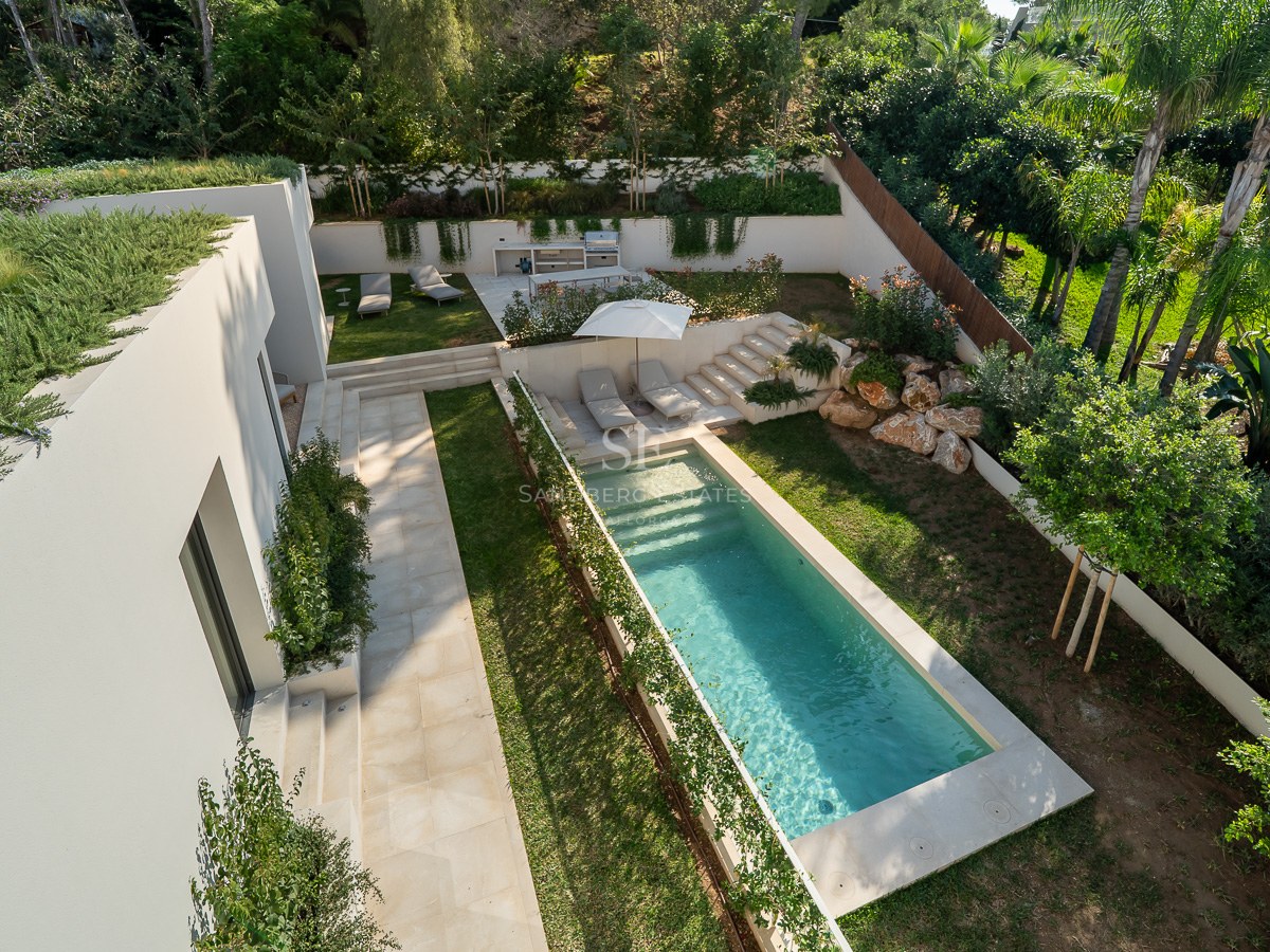 Vue en plongée d'une piscine rectangulaire turquoise entourée de jardins luxuriants et d'une villa blanche moderne.