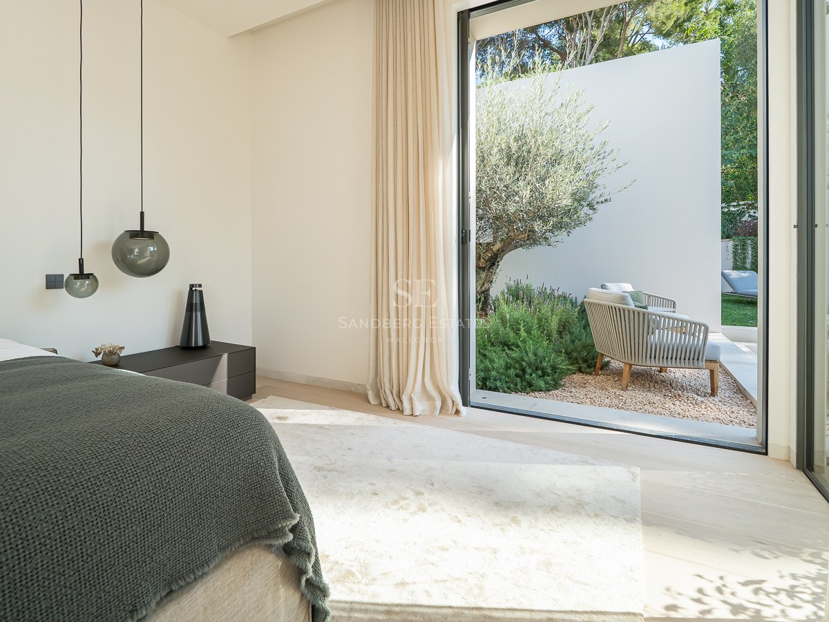 Chambre moderne avec un grand lit, des suspensions design et des baies vitrées ouvrant sur un jardin avec un olivier.