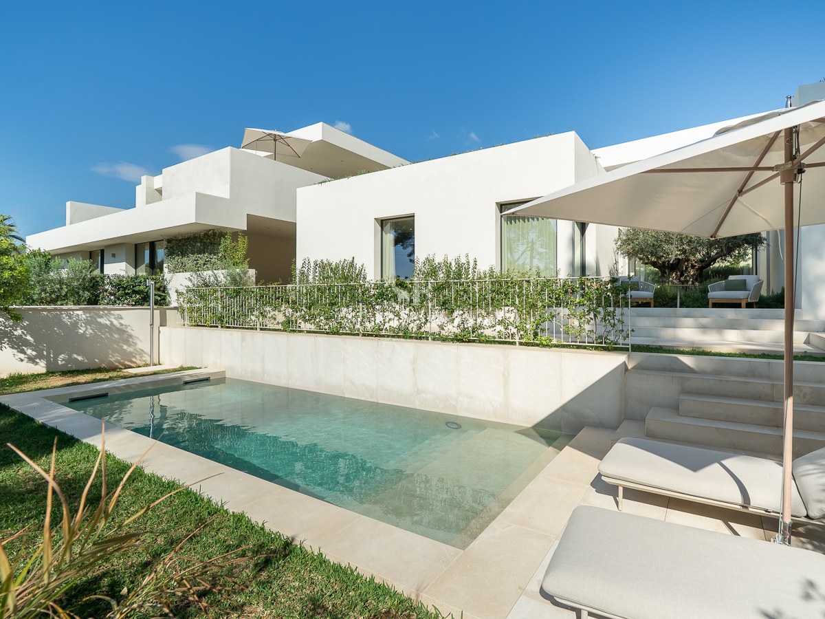 Villa blanche moderne avec piscine rectangulaire, patio en pierre, bains de soleil et jardin verdoyant sous un ciel bleu.