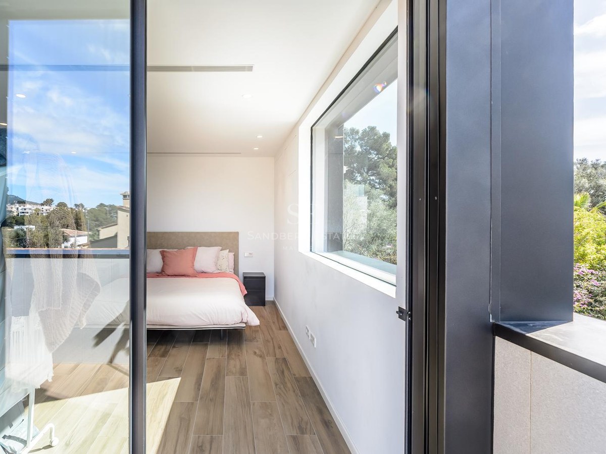 Dormitorio moderno con suelos de madera, una cama con cojines rosas y grandes ventanales con vistas a árboles verdes.
