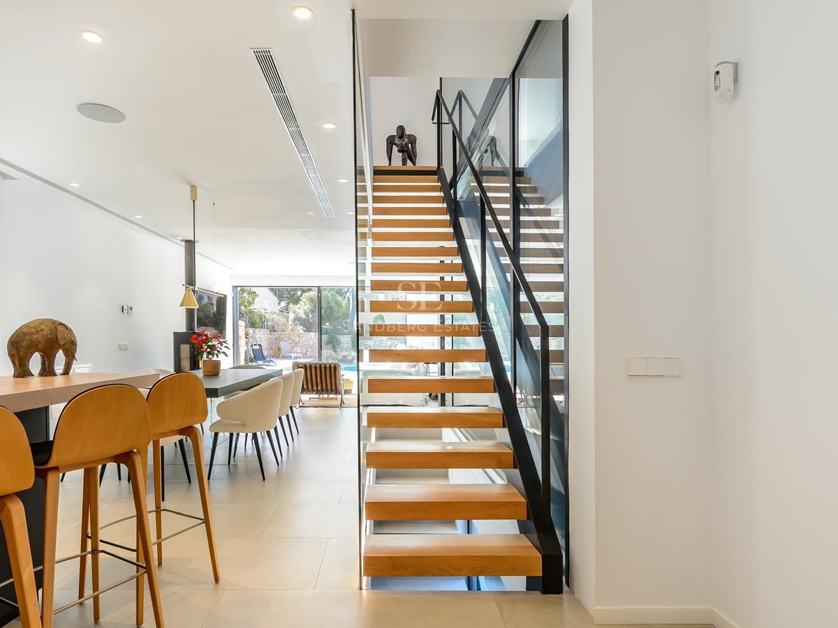 Interior moderno con escalera flotante de madera, barandilla de cristal y salón luminoso al fondo.