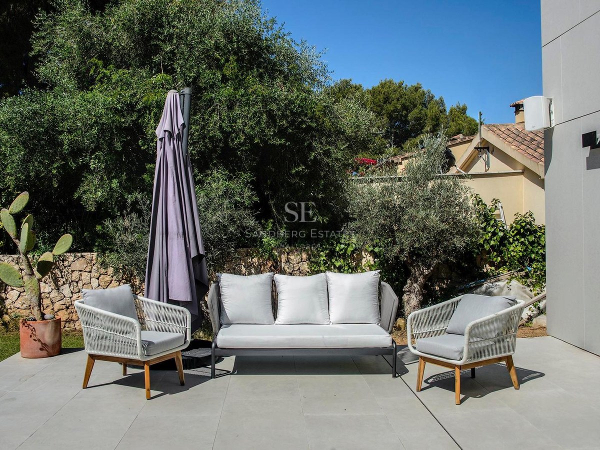 Sofá y sillones de cuerda trenzada gris en terraza de piedra rodeada de vegetación mediterránea bajo cielo azul.