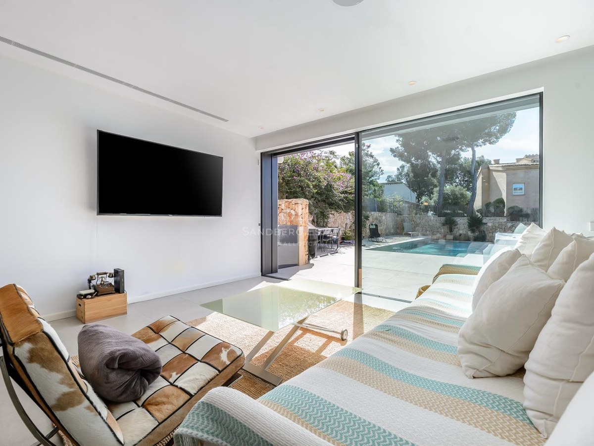 Salón luminoso con sofá a rayas, televisor grande y puertas de cristal que dan a una terraza con piscina.