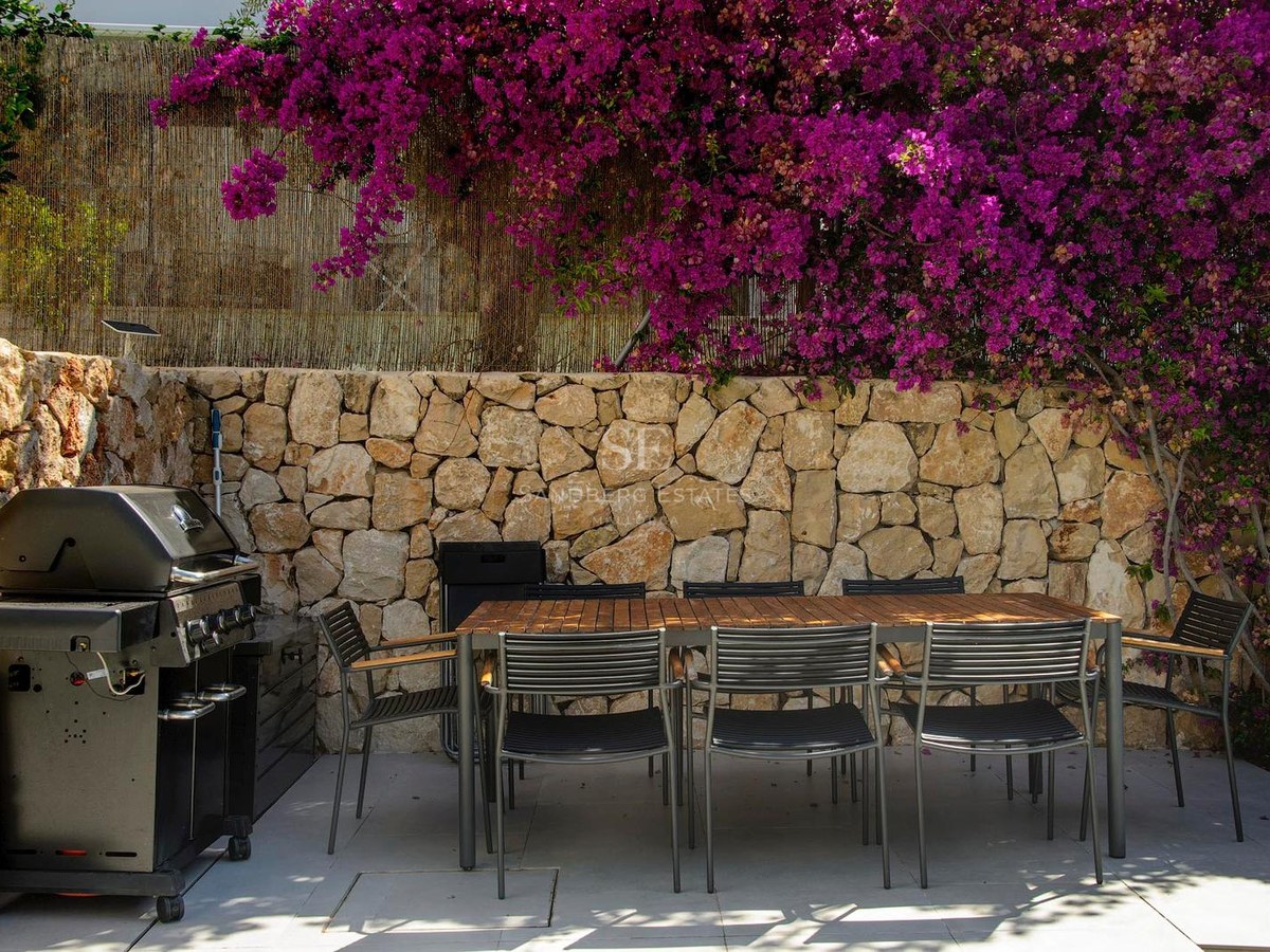 Terraza con mesa de madera, sillas negras, barbacoa y muro de piedra con flores de buganvilla rosa.