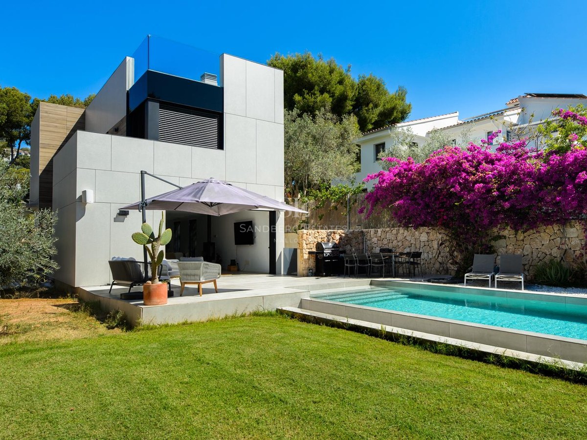Exterior de villa moderna con piscina azul, césped verde y buganvilla morada contra un muro de piedra.