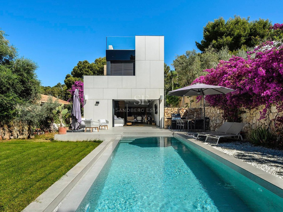 Villa blanca moderna con una piscina rectangular, césped verde y buganvillas púrpuras bajo un cielo azul despejado.
