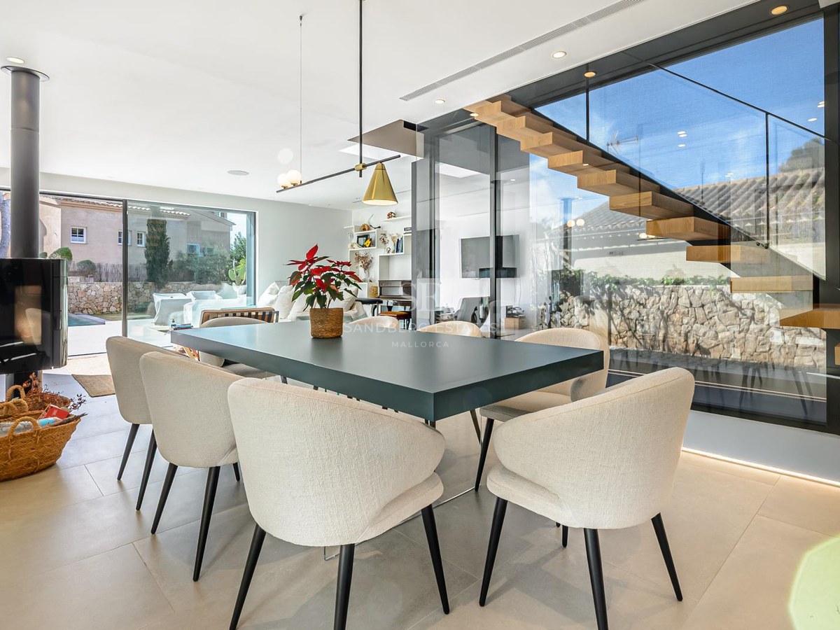 Comedor contemporáneo con mesa oscura, sillas beige, escalera de madera flotante y grandes ventanales de cristal.