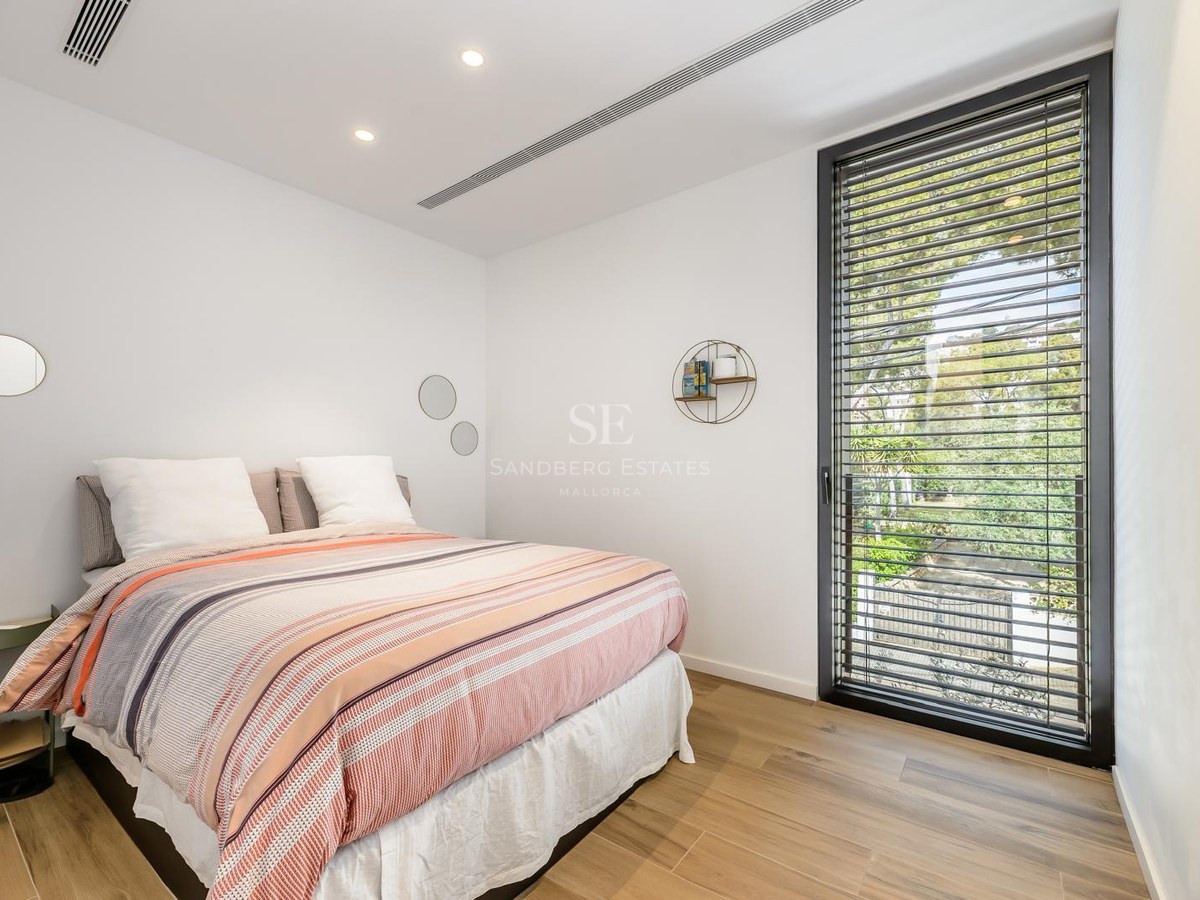 Dormitorio luminoso con cama a rayas, suelos de madera y un gran ventanal con persianas que da a árboles verdes.