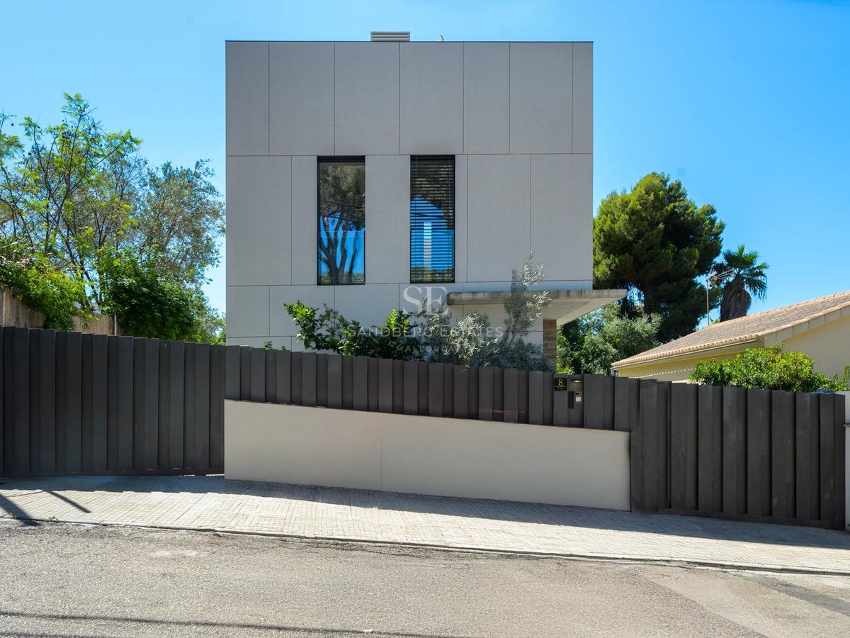 Vista exterior de una casa cúbica blanca moderna con una valla metálica gris oscuro y árboles verdes bajo un cielo azul.