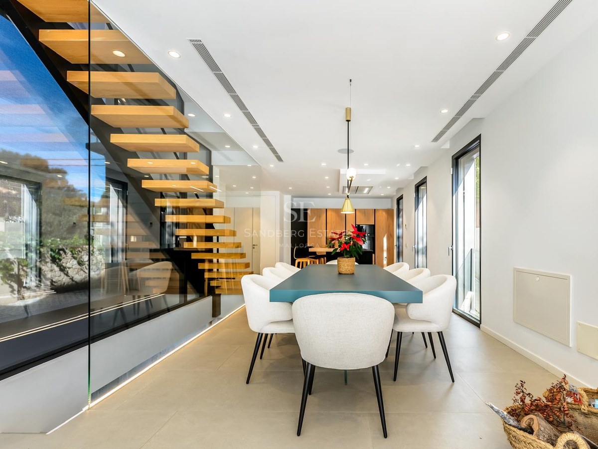 Comedor moderno con mesa azul, sillas blancas y una escalera de madera flotante tras un panel de cristal.