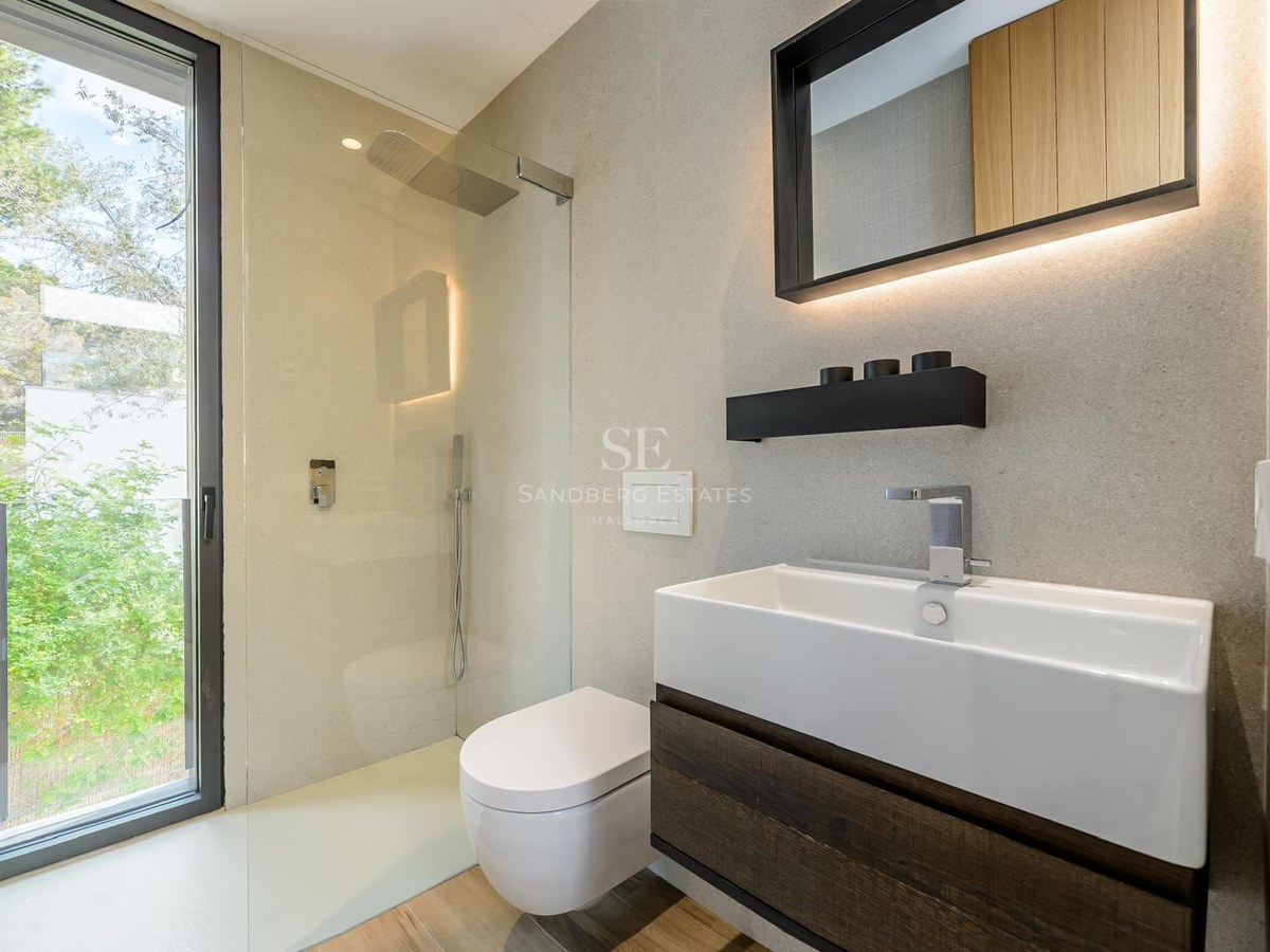 Baño moderno con lavabo blanco, mueble de madera oscura, ducha de cristal y ventanal con vistas al jardín.