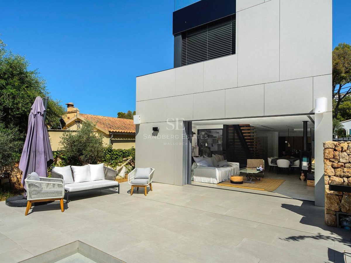 Terraza exterior moderna con muebles blancos junto a la zona de estar abierta de una villa y una pared de piedra.