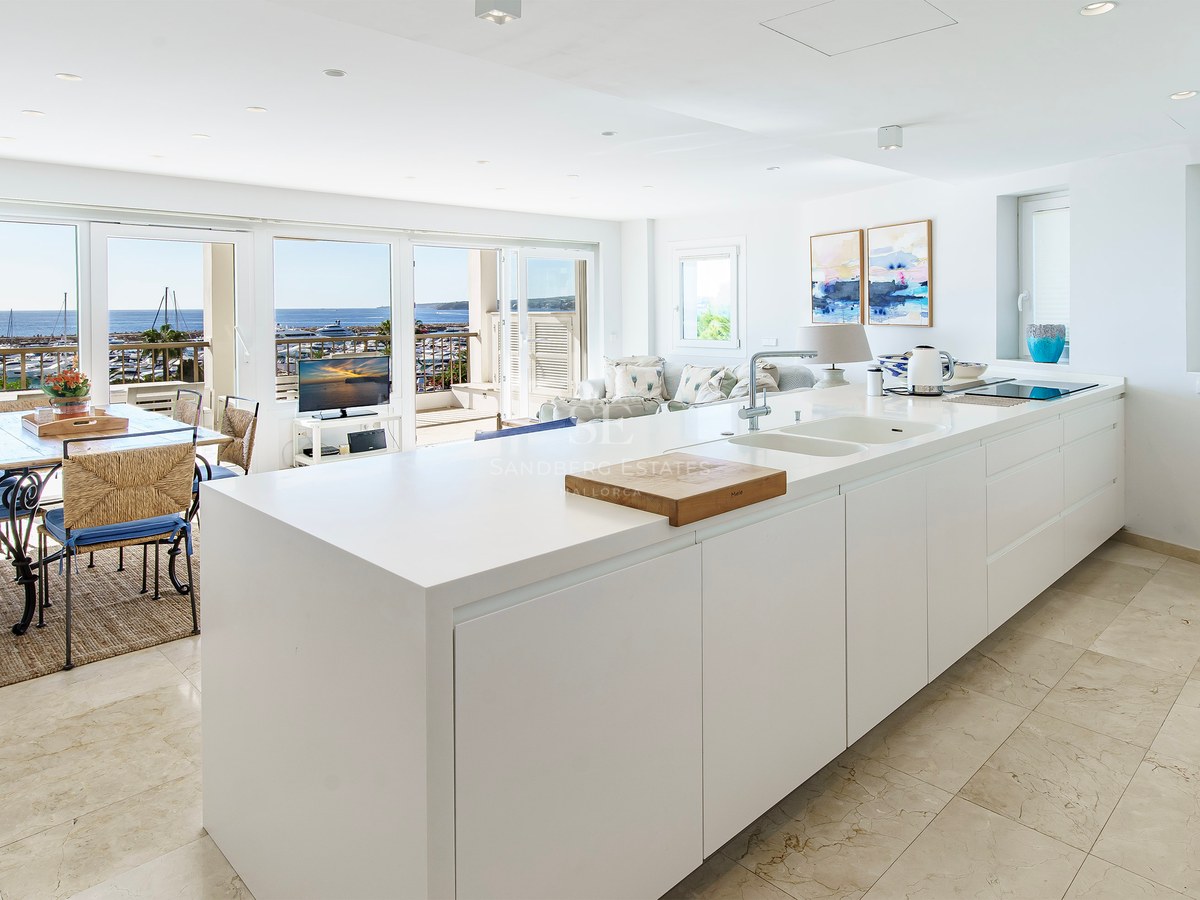 Minimalistisk vit köksö som leder till en matplats med panoramautsikt över marinan och havet.