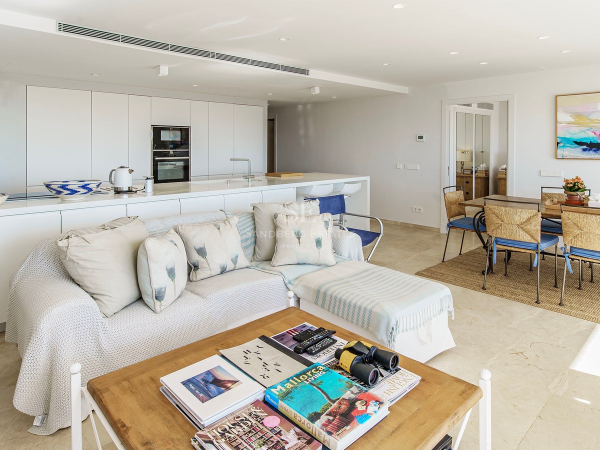 Ljust vardagsrum med öppen planlösning, vit soffa, träbord, matgrupp och minimalistiskt kök i bakgrunden.