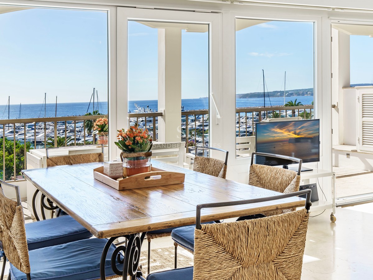 Soligt matrum med träbord och korgstolar med utsikt över en marina fylld med yachter och det blå havet.