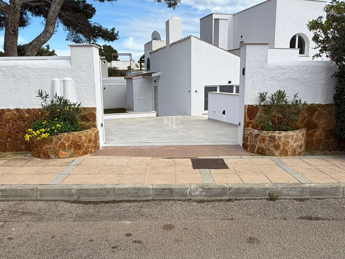 Entré till en vit villa med stenmurar, planteringar med gula blommor och stenlagd uppfart under tallar.