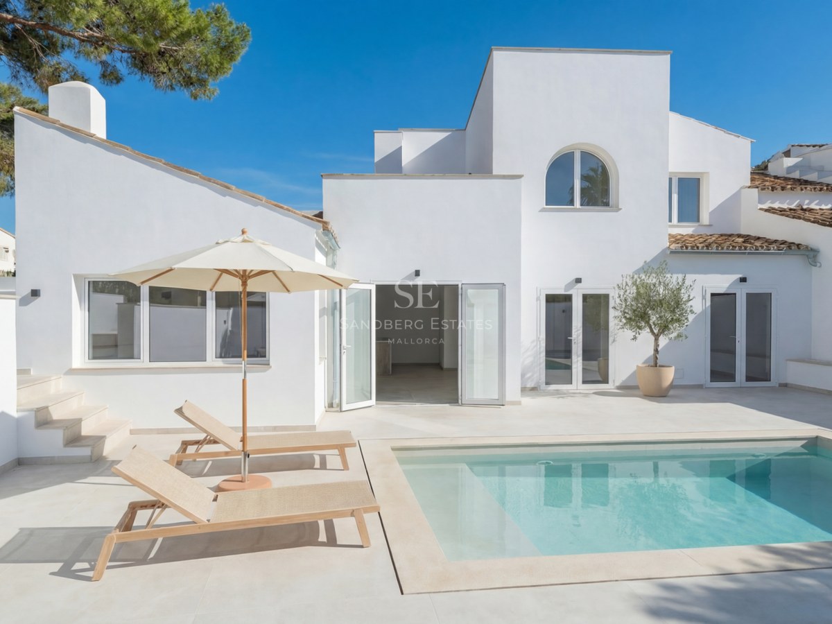 Vit modern villa med rektangulär pool, ljus stenterrass och solstolar under en klarblå himmel.