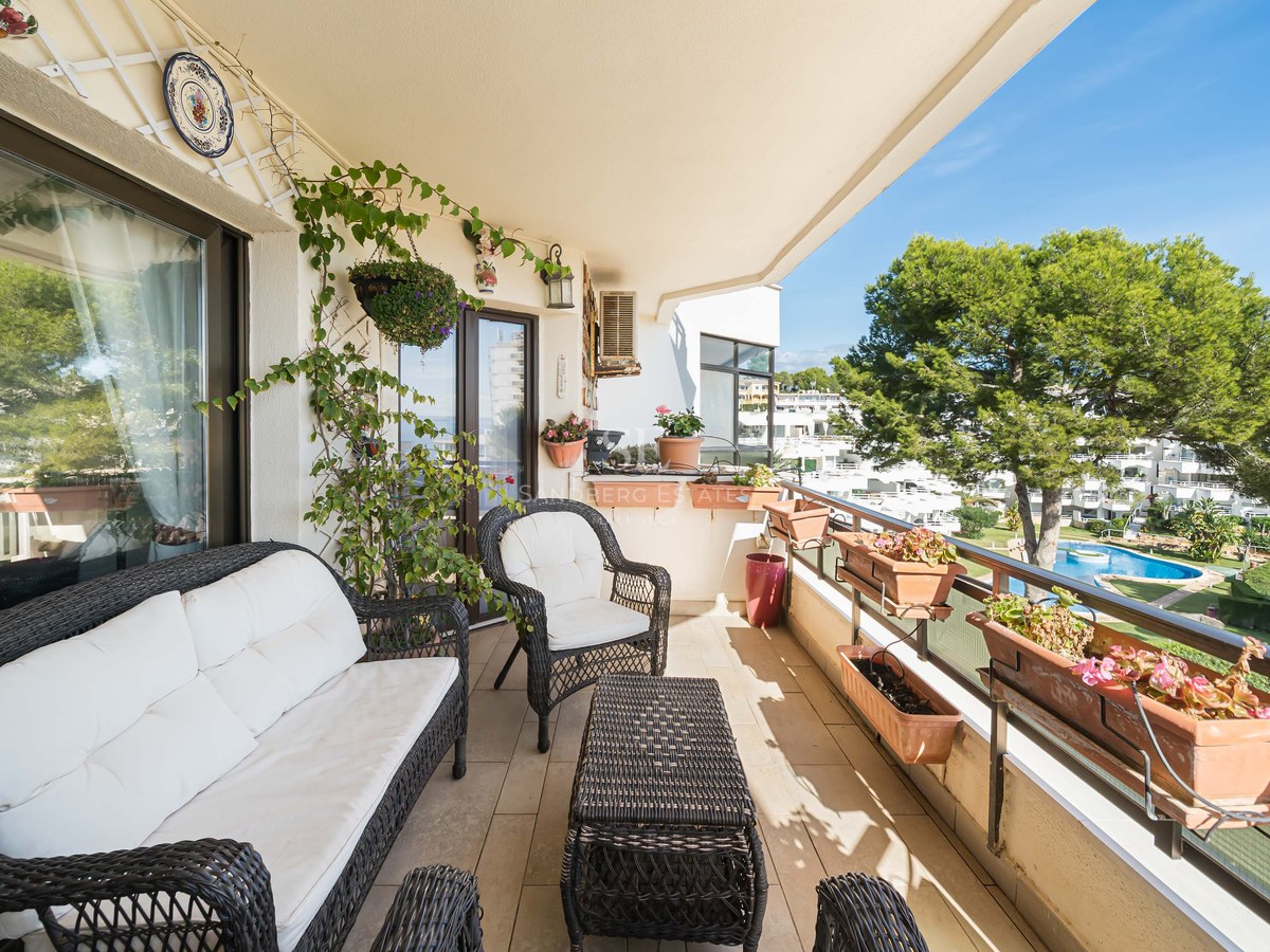 Balcony with dark wicker furniture, white cushions, and many potted plants overlooking a swimming pool and garden.