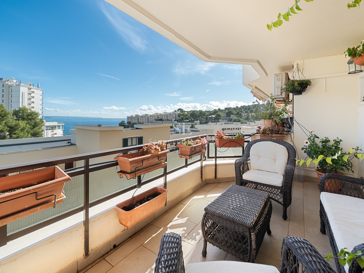 Balcony with wicker furniture, white cushions, and planters overlooking a blue sea and coastal buildings.