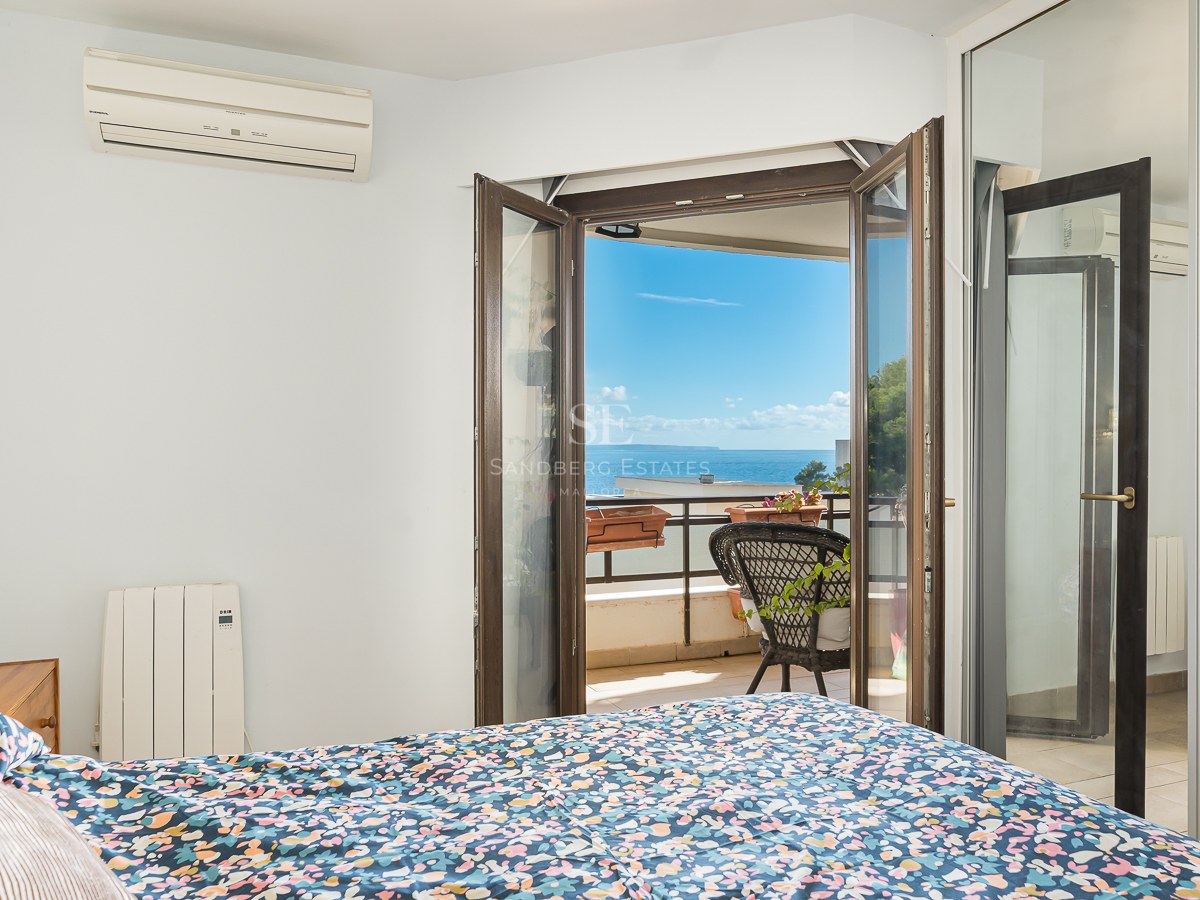 Bedroom with a floral bed overlooking a balcony with a black wicker chair and a wide view of the blue ocean.