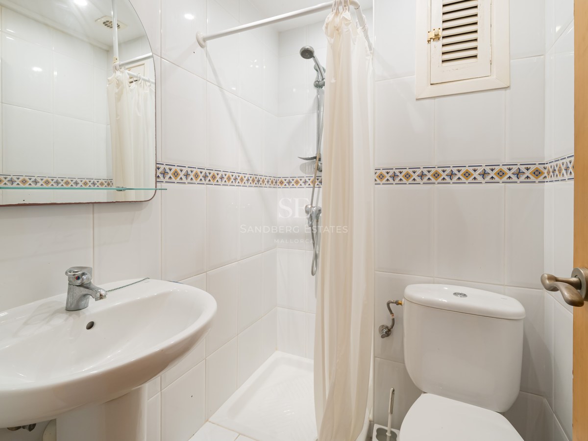 Bright bathroom with white tiles, a pedestal sink, toilet, and a walk-in shower with a white curtain.