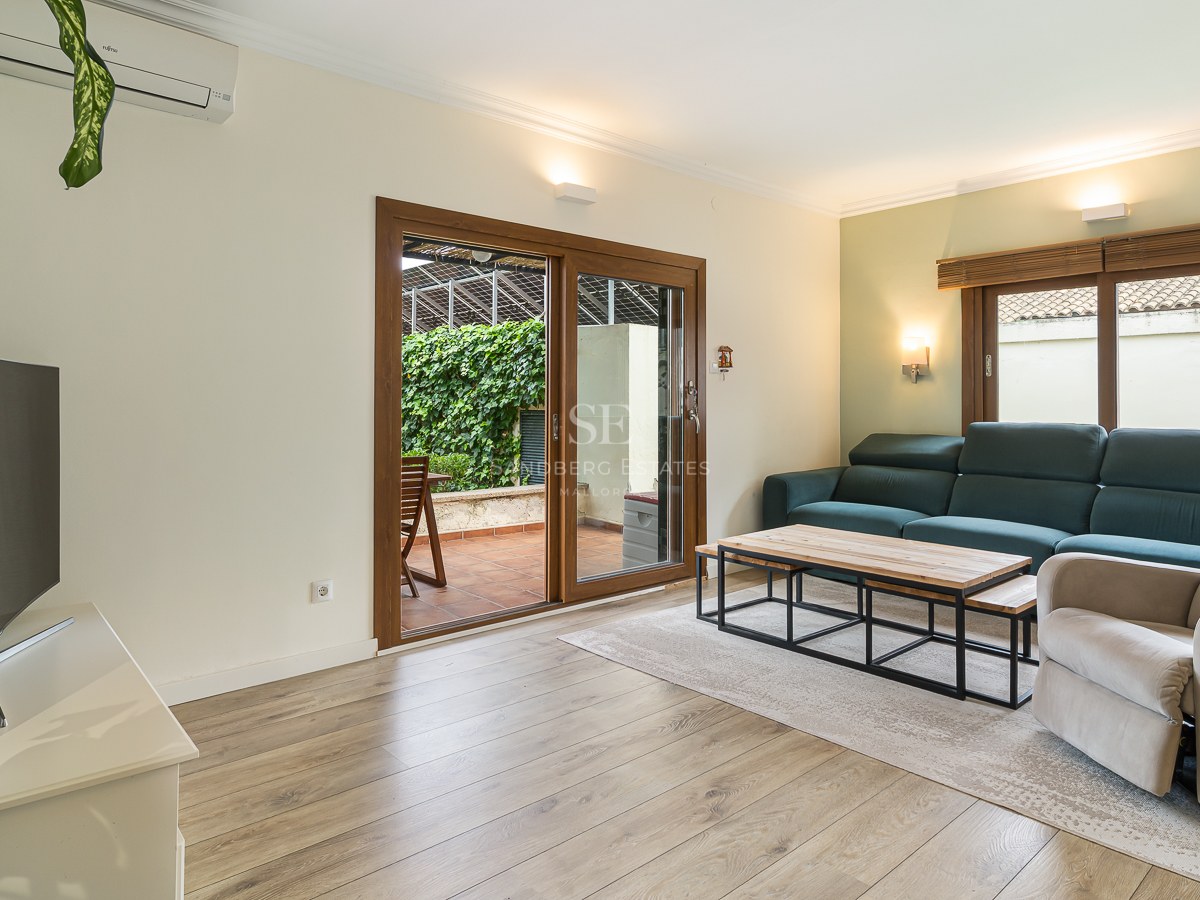 Bright living room with teal sofa, armchair, wood floors, and glass doors leading to a lush green terrace.