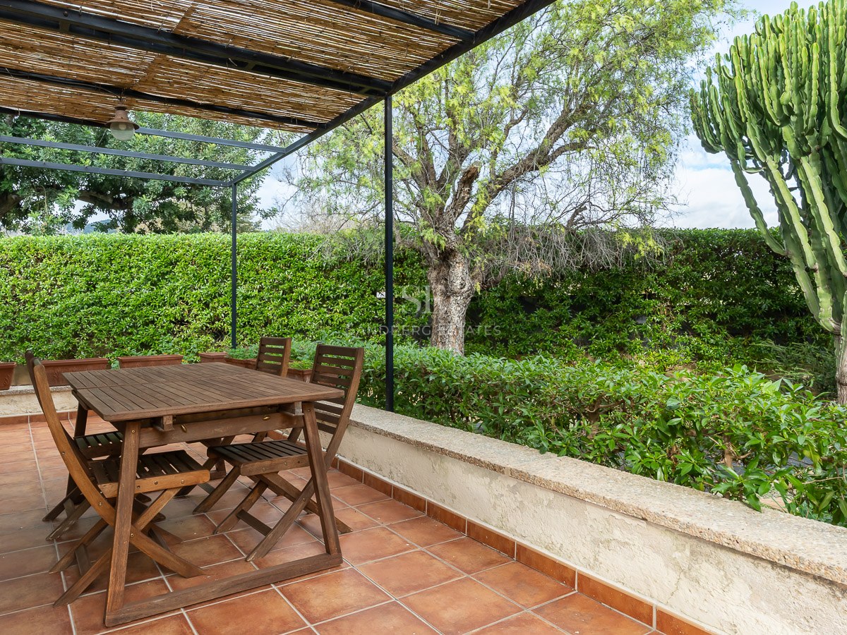 Wooden dining set under a reed pergola on a terracotta tiled terrace surrounded by lush greenery and a large cactus.