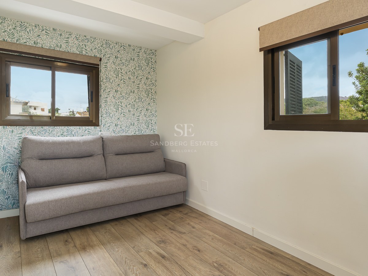 Modern bedroom featuring a grey sofa, botanical leaf pattern wallpaper, wooden floors, and natural light from two windows.