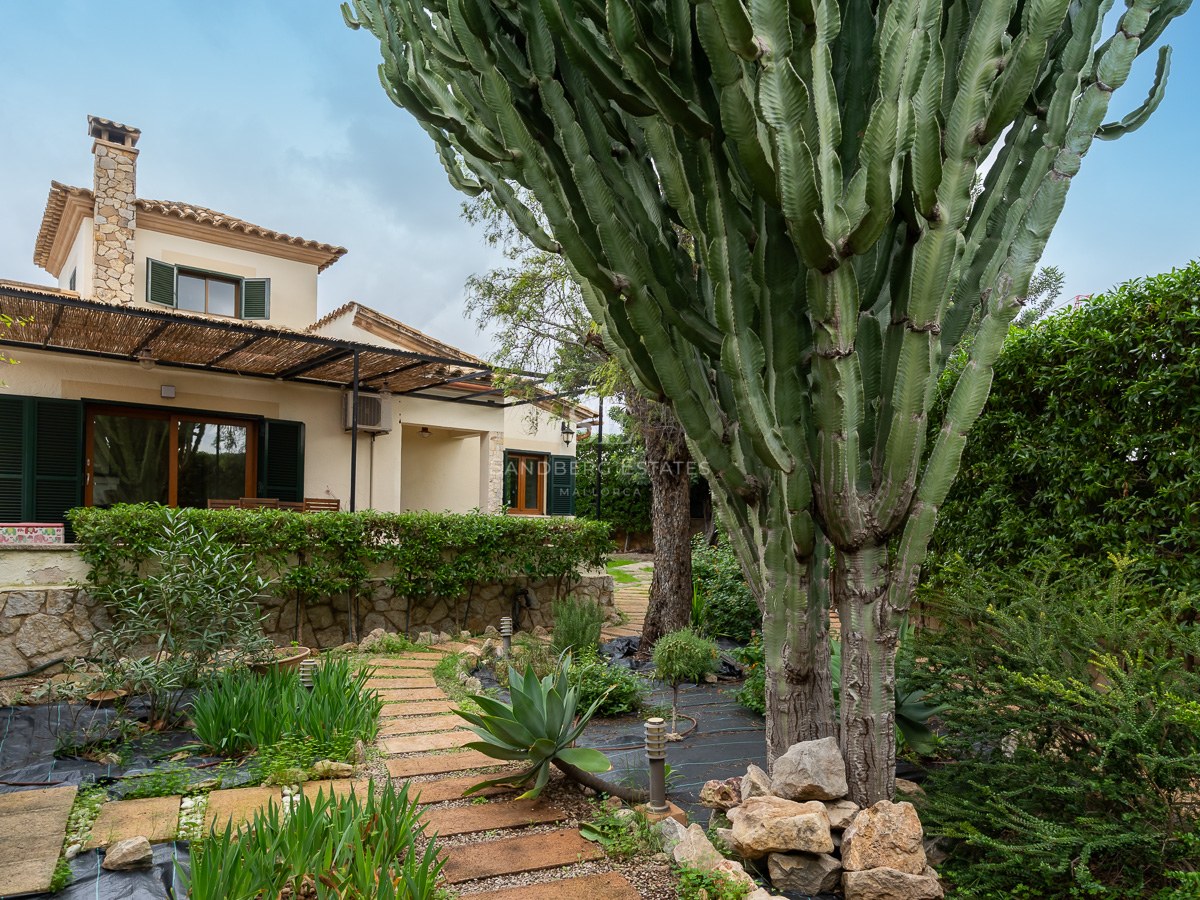 View of a lush garden featuring a large cactus, stone pathway, and a Mediterranean home with green shutters.