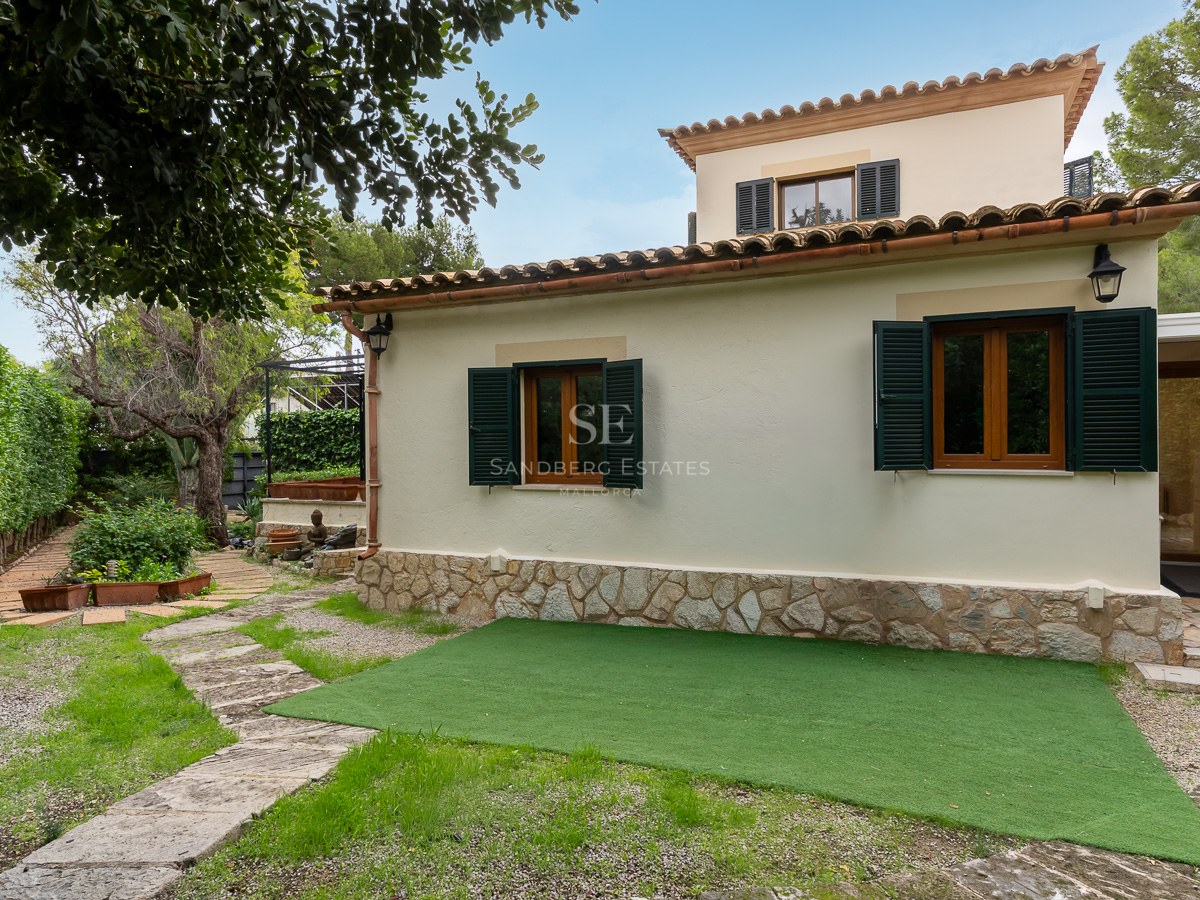 Cream stucco villa with green shutters, stone base, and a winding stone garden path under a clear blue sky.