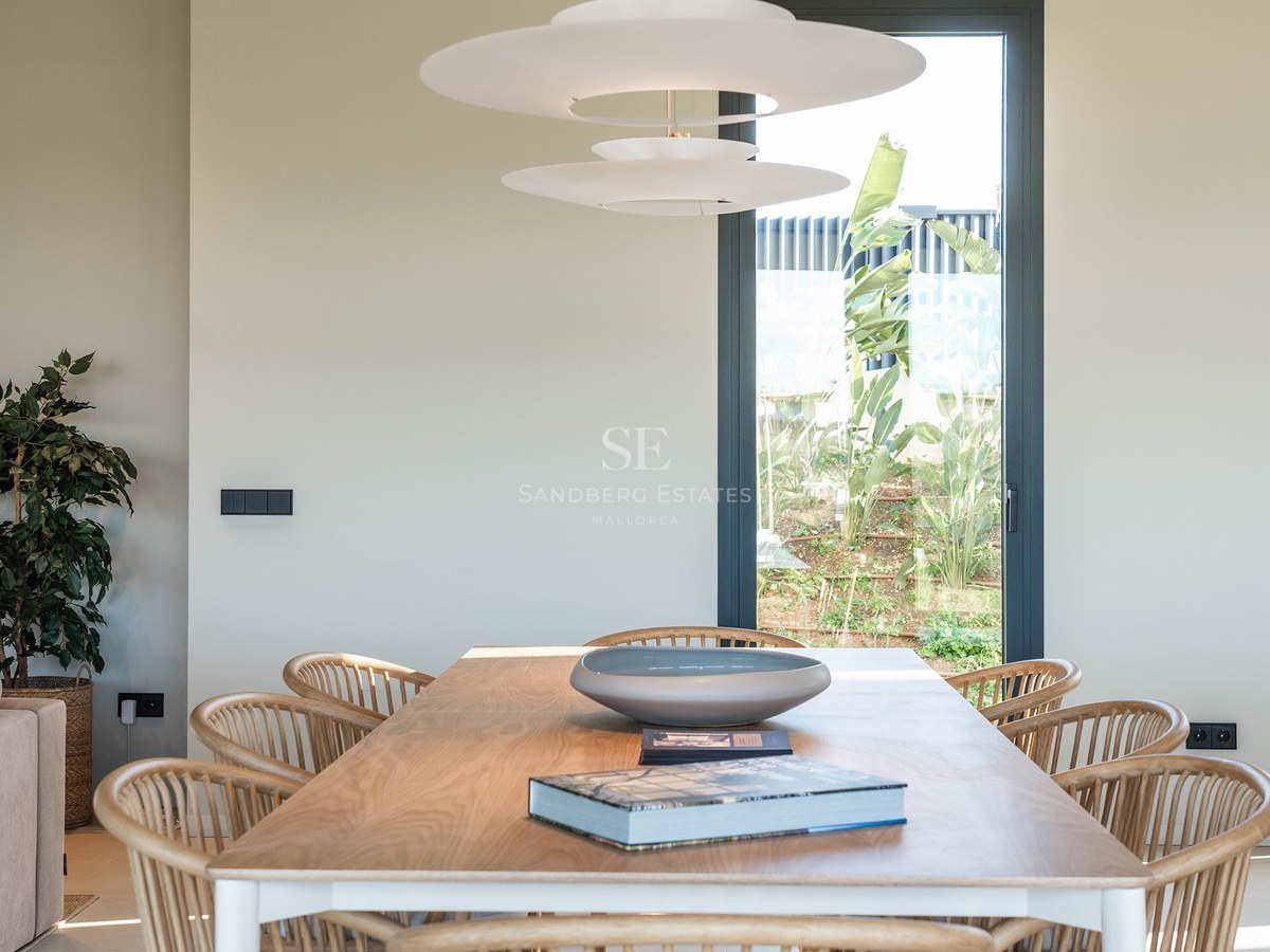 Modern dining room featuring a wooden table, rattan chairs, a designer pendant light, and a window with garden views.