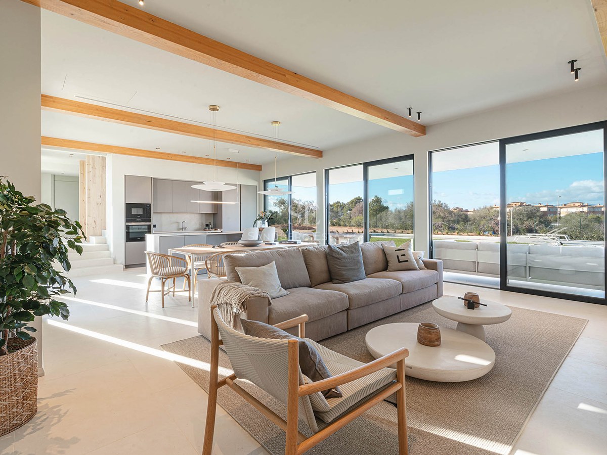 Spacious modern living room with large beige sofa, wooden ceiling beams, and floor-to-ceiling glass doors.
