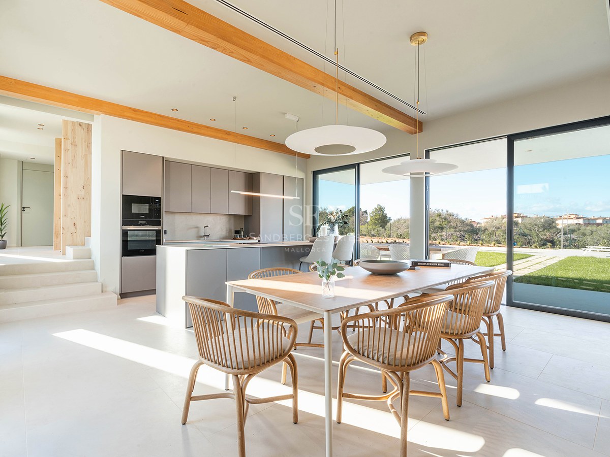 Bright dining room with wooden table and chairs next to a minimalist kitchen with large glass doors to garden.