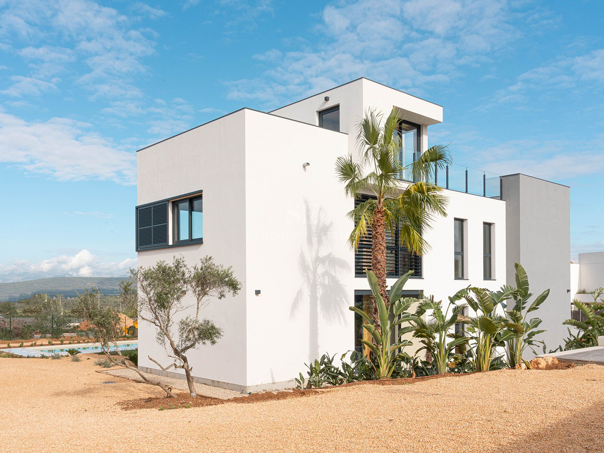 Exterior of a three-level white villa with modern architecture, palm trees, and a gravel yard under a blue sky.